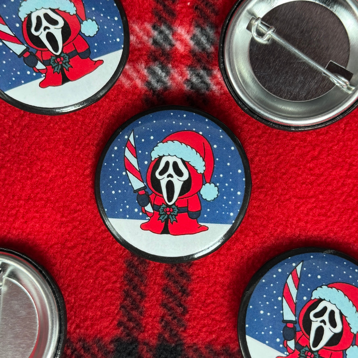 Santa Ghostface pinback buttons on red plaid fabric, showing the masked character holding a candy cane-striped knife with a Santa hat in the snow.