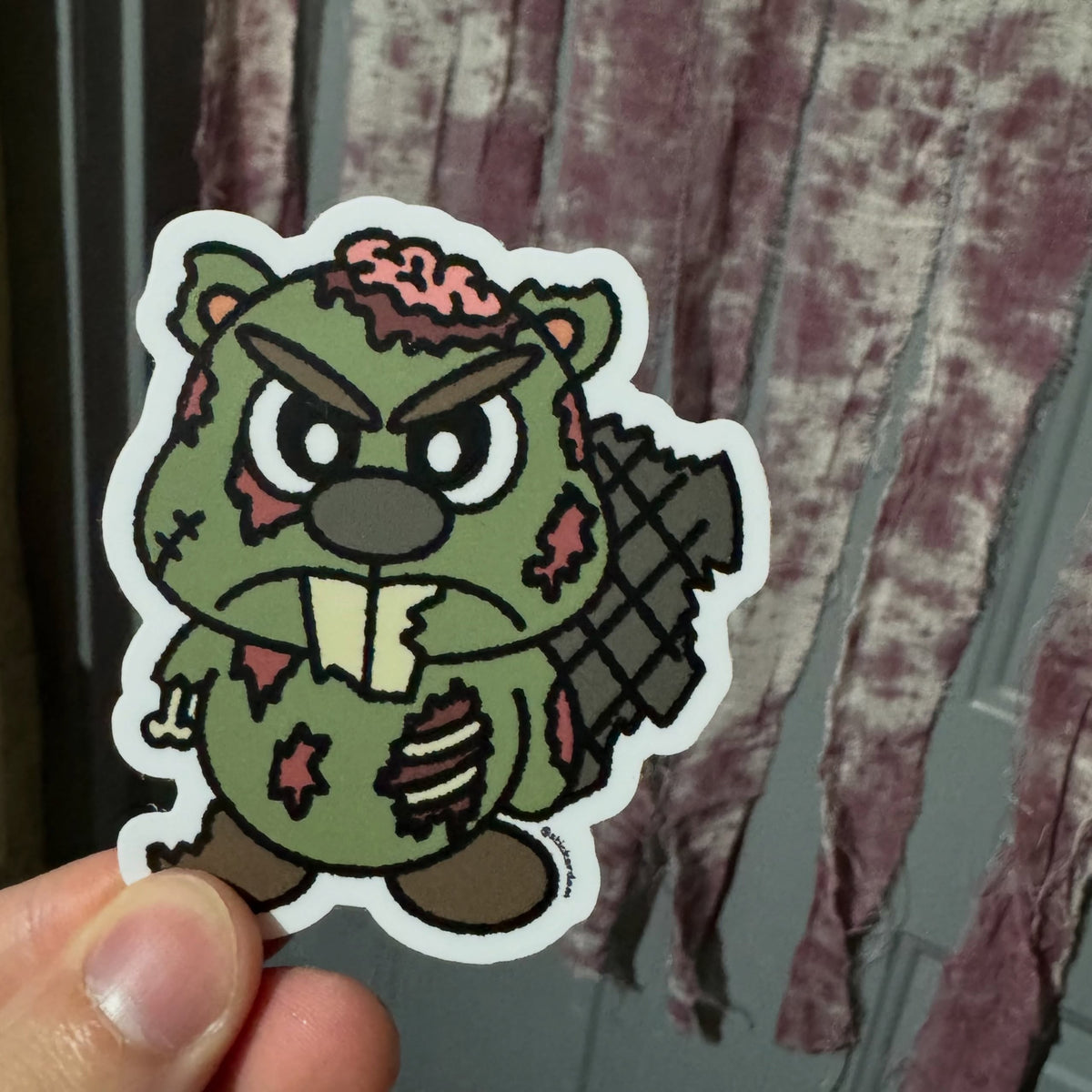 Hand holding a Zombie Dammy zombie beaver vinyl sticker in front of hanging Halloween decorations.