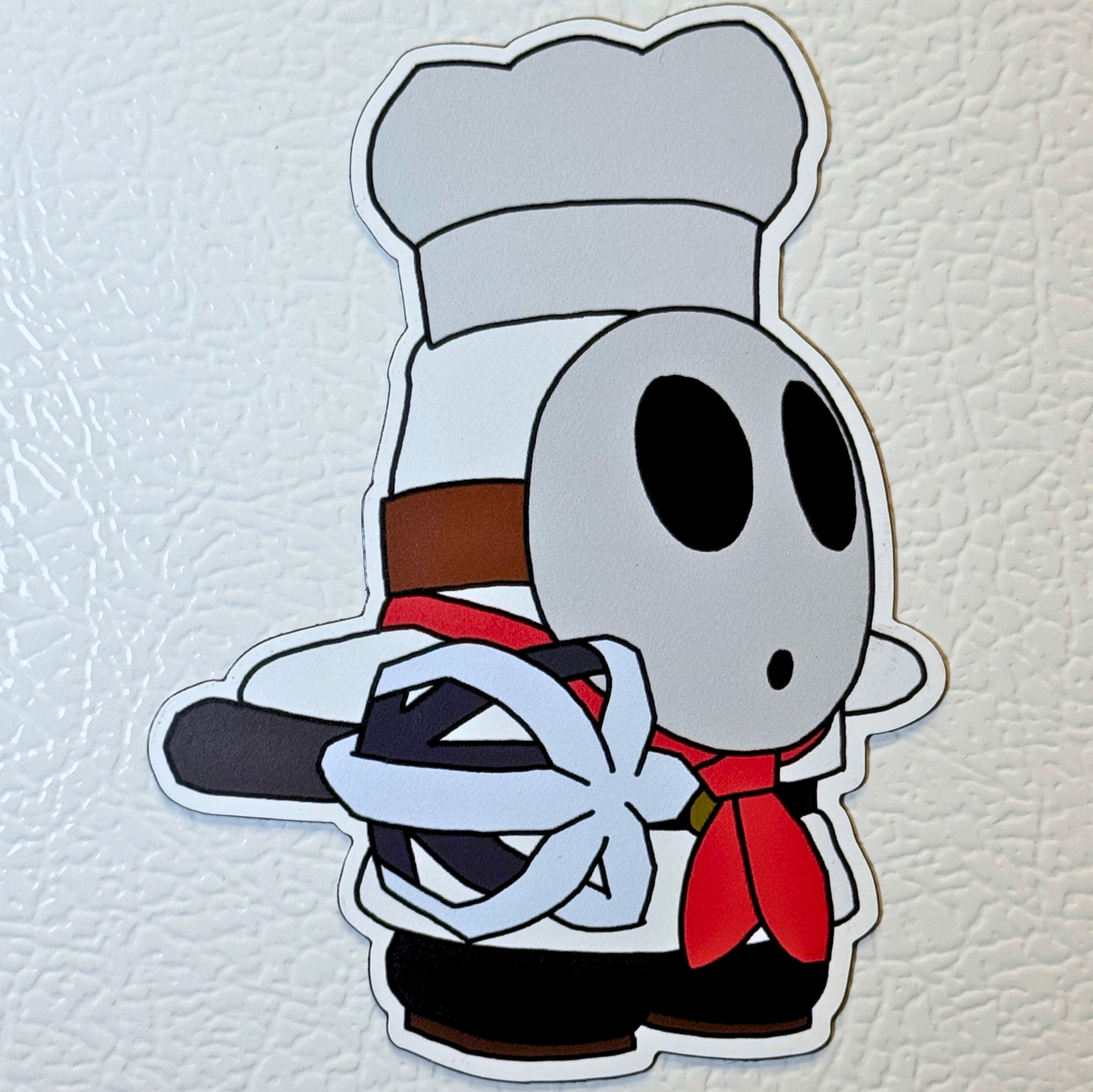 Shy Guy wearing a chef's hat and holding a spatula on a textured refrigerator 