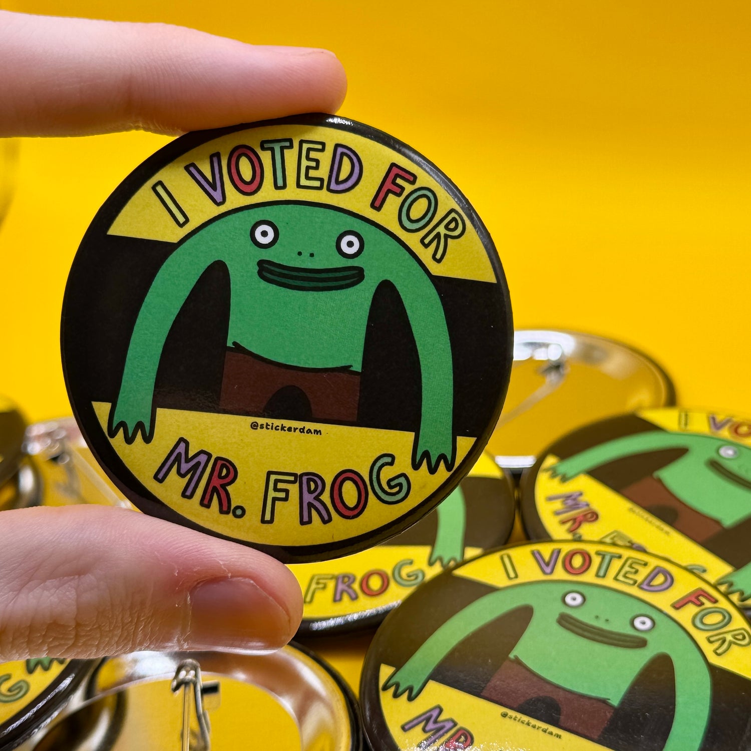 Button badge with 'I Voted For Mr. Frog' design inspired by Smiling Friends held by a hand against a yellow background