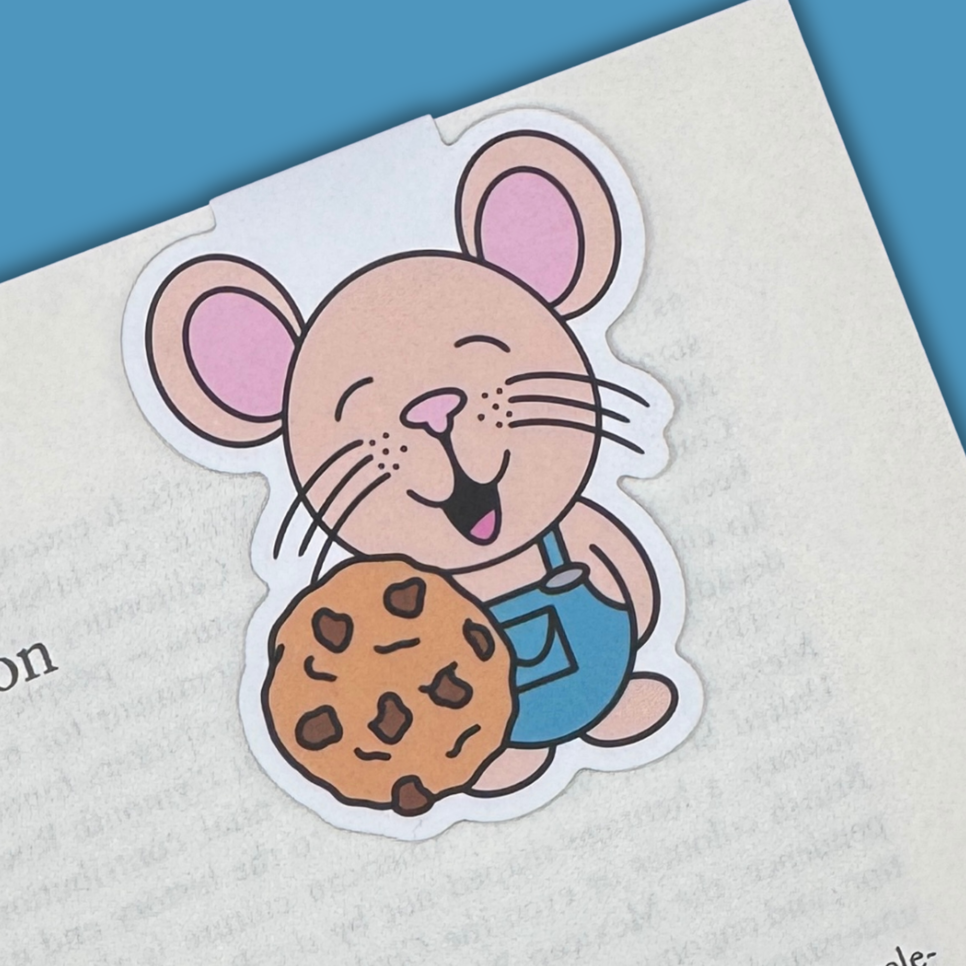 Cartoon mouse bookmark holding a chocolate chip cookie in a book with a blue background