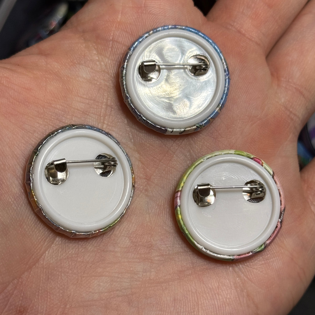 Three upcycled Pokémon card buttons resting face down in a palm, showing white backs and metal pin clasps.