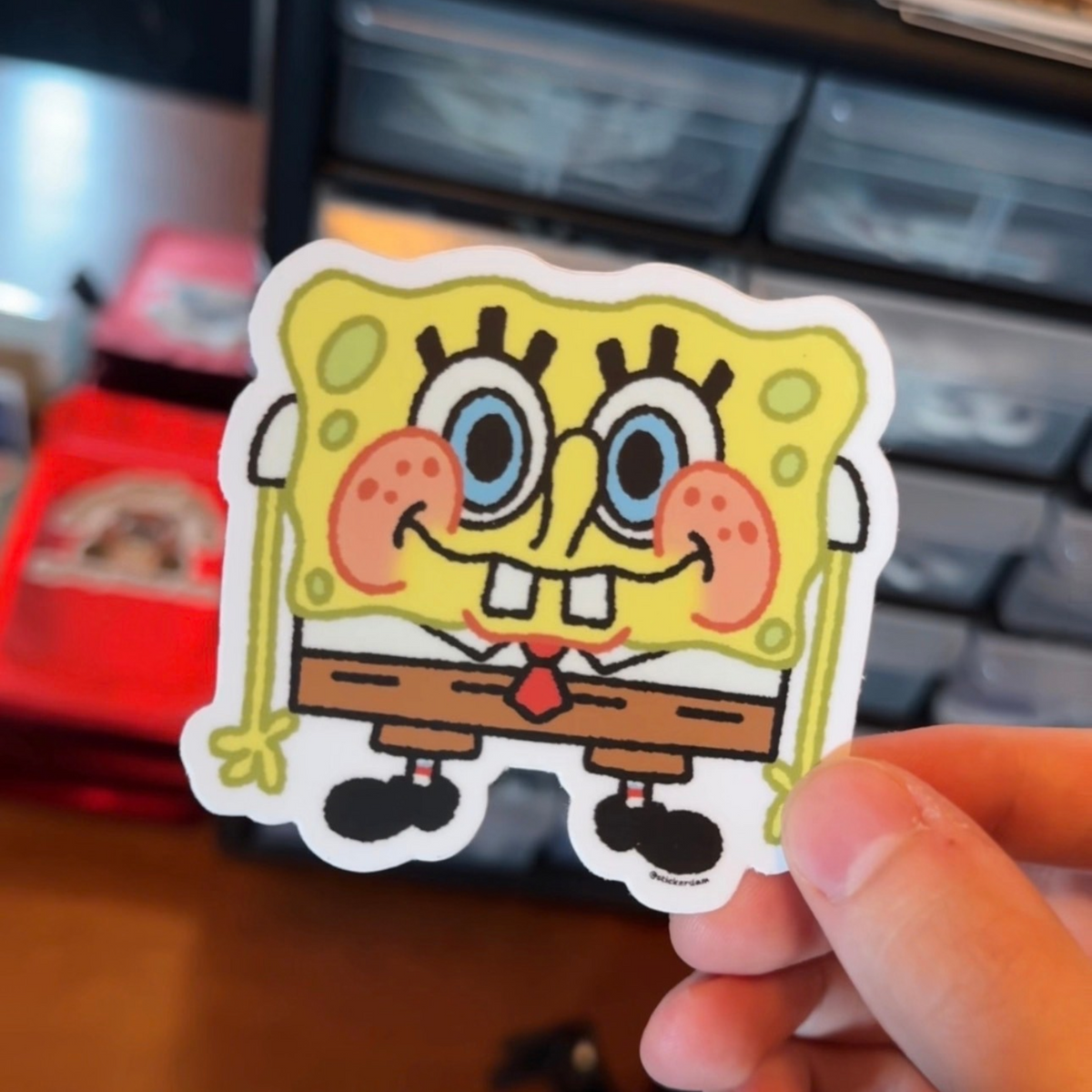 Hand holding a white-bordered SpongeBob-inspired fan art sticker with big blue eyes, rosy cheeks, and brown shorts.